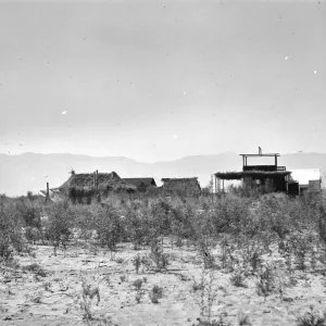 Black and white photograph of a group of buildings in a desert setting with brush in the foreground and hills in the distance.