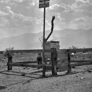 Black and white photograph of a signpost above a wooden fence that has signs to Blackwater, Wildrose, Tanks, Ash Meadow, Tule Hole, Bennetts Wells, Wondermine, and Salt Creek. There are mountains in the distance.