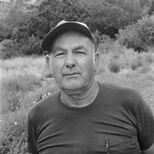 Black and white photograph of a man wearing a short sleeved shirt and a baseball cap, looking at the camera in a garden setting.