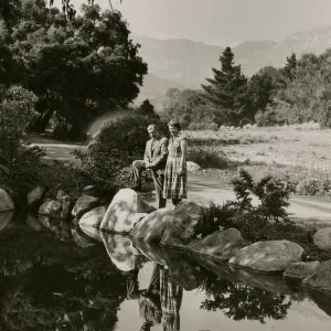 Black and white photo of a man in a suit and a woman in a plaid dress look out over a pond lined with large rocks, the man has a leg propped up on one of the rocks. 