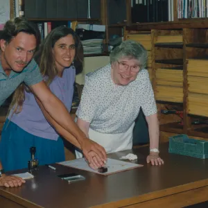 A man in grey, a woman with long hair wearing in a lavender shirt, and an older woman in white look toward the camera while standing in front of shelves lined with many binders, books, and yellow memo pads. The three lean forward over a desk where they put their hands together, applying a red stamp to a document sitting on the desk. .