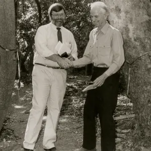 A black and white photo of two men, one is bearded, wearing glasses and a light-colored dress shirt and dark tie, the other has light hair and wears dark slacks. Both are holding documents and stand in front of a cut ceremonial ribbon tied between two trees. A sign reading "Easton Tr" is cutoff at the edge of the image. 
