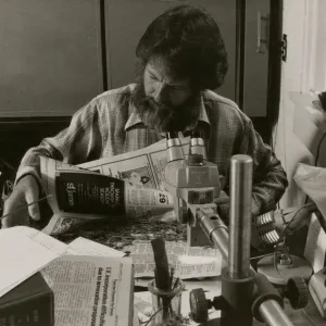 A black and white photo of a bearded man in a plaid shirt looking through newspapers while sitting at a desk cluttered with newspapers, books, and scientific instruments.