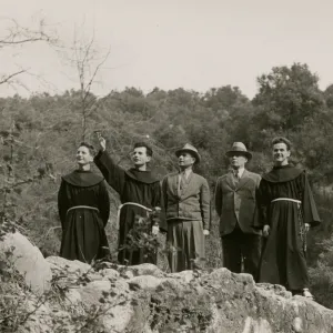 A black and white photo of five men look into the distance while standing on a rocky bluff. Three of the men wear robes ties with ropes at the waist and two wear suits and trilby hats. One of the robbed men points out in the distance where the group is looking.