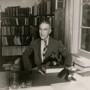 A black and white photo of a man in a suit and plaid tie sits at a desk in front of shelves full of books. 