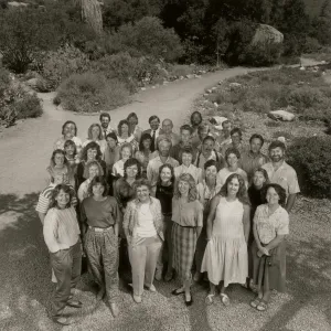 A black and white photo of a group of 36 smiling people taken from a high angle. They stand in front of a gravel road lined with stones and rough shrubbery.