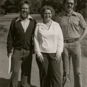 A black and white photo of two men and a woman, smiling with windswept hair, in a rocky field at the edge of a copse of trees. 