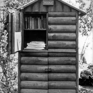 Black and white photograph of log cabinet labeled books with one door open and books inside in wooded area. 