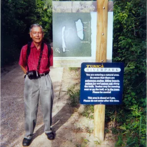 Photograph of male wearing red shirt and camera standing at trail sign with lush green background. 