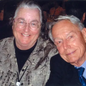 Flash photograph of female wearing black grey shirt with glasses and male wearing black suit blue tie both at event smiling. 