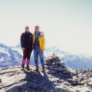 Photograph of male wearing yellow jacket and female wearing black and pink clothing standing on top of mountains with rocky and snowy terrain.
