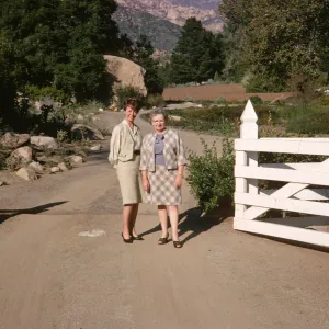 Two women, one older and one younger, in pale colored suits stand, smiling on a gravel road in an open white fence gate. The road behind them is lined with rocks and trees, with mountains and a large boulder visible in the background.