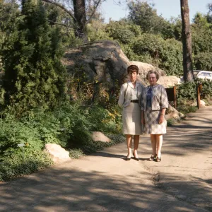 Two women with serious expressions wearing pale colored suits stand on an uneven paved road. Next to them at the edge of the road there is an evergreen tree among thick greenery, and behind them are a few large boulders and 2 pale colored, older model cars. 