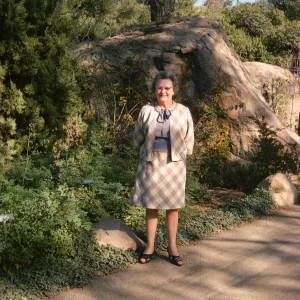 An older woman in a pale suit smiles at the edge of a paved road. Next to her at the edge of the road is a tall evergreen tree surrounded by low greenery and behind her is a large boulder.