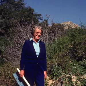 An older woman dressed in royal blue smiles in front of a rocky hill coated in short vegetation and bare-branched bushes. A rolled blueprint is held in her right hand. 