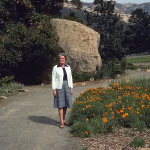 An older woman in a grey plaid shirt and white cardigan stands on a curving, gravel road next to a field of orange flowers. A large boulder sits among a copse of dark green trees behind her, on the other side of the road. 