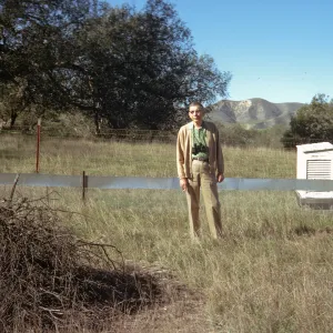 A man khaki colored pants and sweater with a set of binoculars around his neck stands in a field crossed by wire fencing. A large pile of sticks is on the ground to his right, and a white, wooden box  is behind, to his left. 