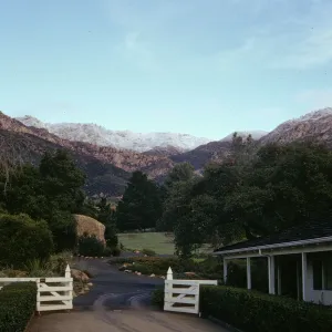 A dark, gravel road lined by neatly trimmed, squared hedges passes by a white, single-story building with a dark, shingle roof and past an open, white fence. The road continues past the fence, curving between bushes, trees, and two fields of flowers and grasses, and a large boulder. Rocky mountains dominate the background. 