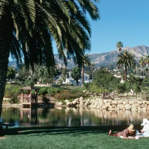 A few people lounge on a lawn beside a large pond. A brown gazebo and many plants behind a stone retaining wall are visible on the opposite side of the pond. 